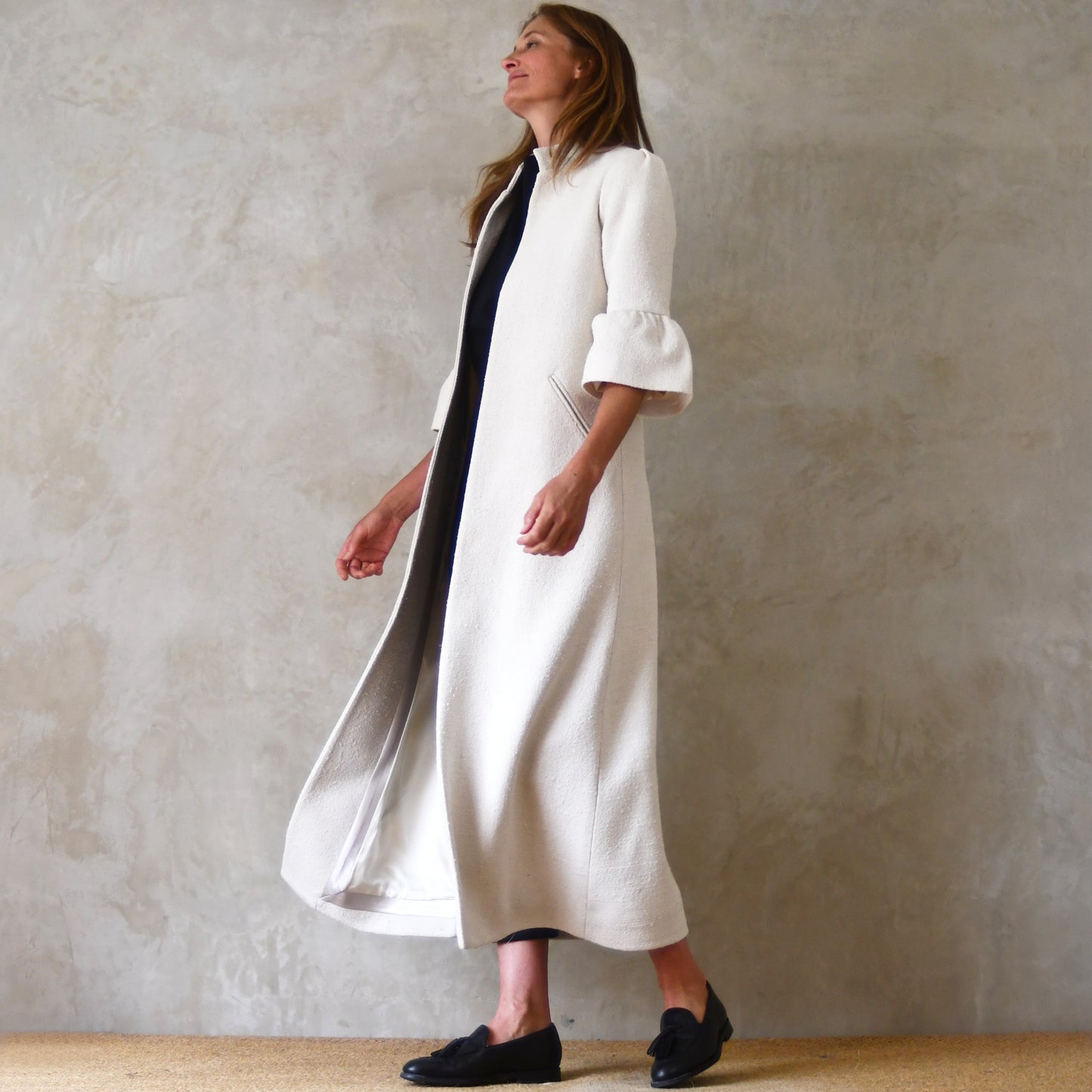 Long White Silk Coat With Bell Sleeves. An A Line Shape elegant coat in pure silk. Nehru collar, gathered cuffs on sleeves and slanted pockets to front. Handmade in Britain. Bespoke options available. Full View.