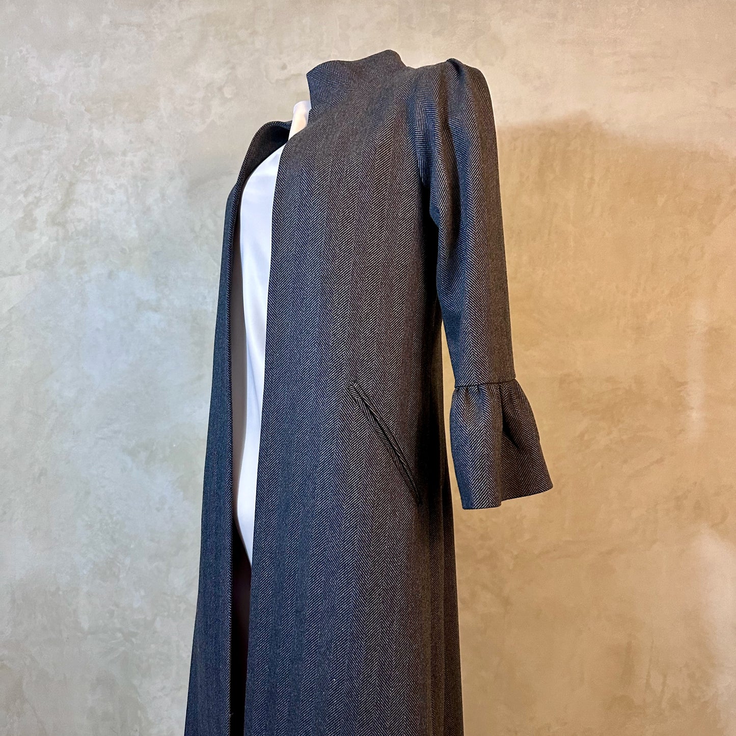 Bell Sleeve A-Line Coat in Charcoal Cashmere Mix Flannel. Bespoke longer length deep grey-coloured A-line coat with gathered shoulder detail, frill cuffs, two slanted pockets to the front and a luscious pink silk lining. Handmade in Britain. Woven in Yorkshire. Side On View.