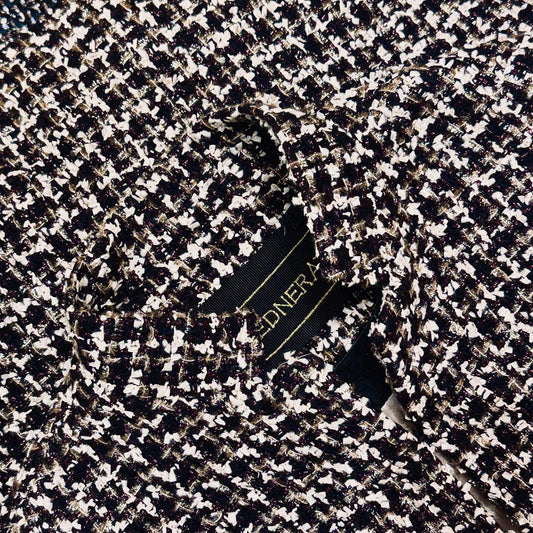 A close-up of a tailored black, white, and metallic tweed fabric jacket with a visible garment label partially showing gold text.