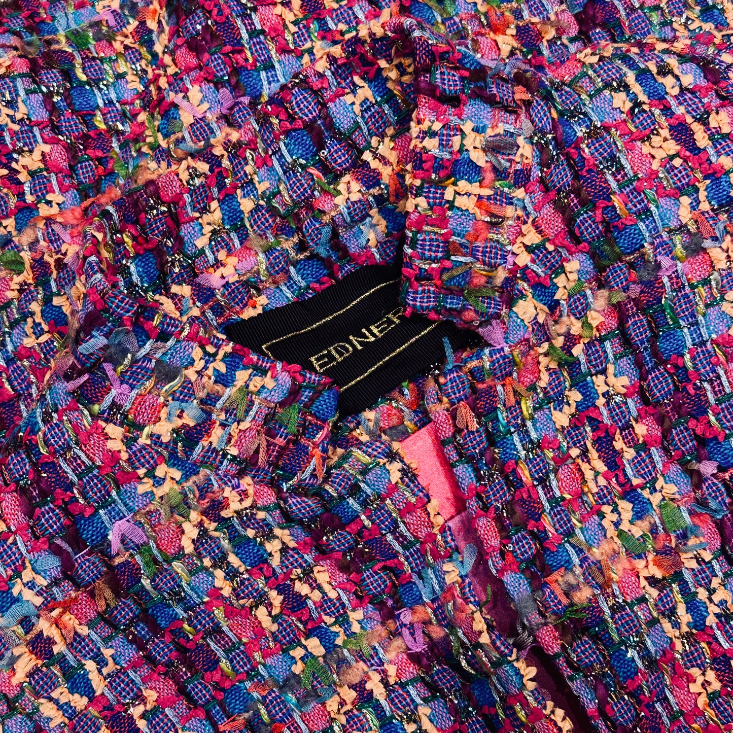A Vintage Style Swing Cape in Raspberry Boucle from Linton Tweeds. Fun, young and elegant. Handmade in Britain. Open View. Close-up of this colourful, textured tweed fabric with vibrant blue, pink, yellow, and red threads. A black label with gold text is partially visible inside this British-made tailored garment.