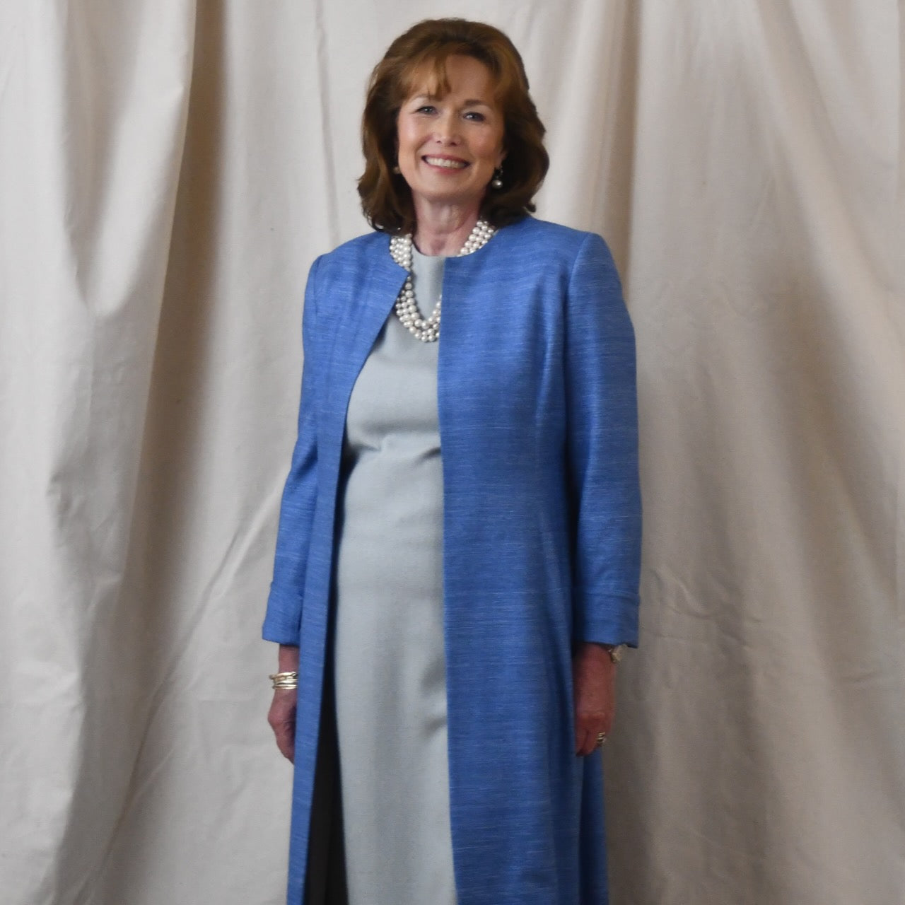 Tailored, Fitted Hourglass shaped coat in a summer-weight Silk Madras in Blue. This is a long-sleeved, calf-length blue summer coat with an hourglass shape. Shown from the side-front. A perfect summer coat for weddings and special occasions. Handmade in Britain.
