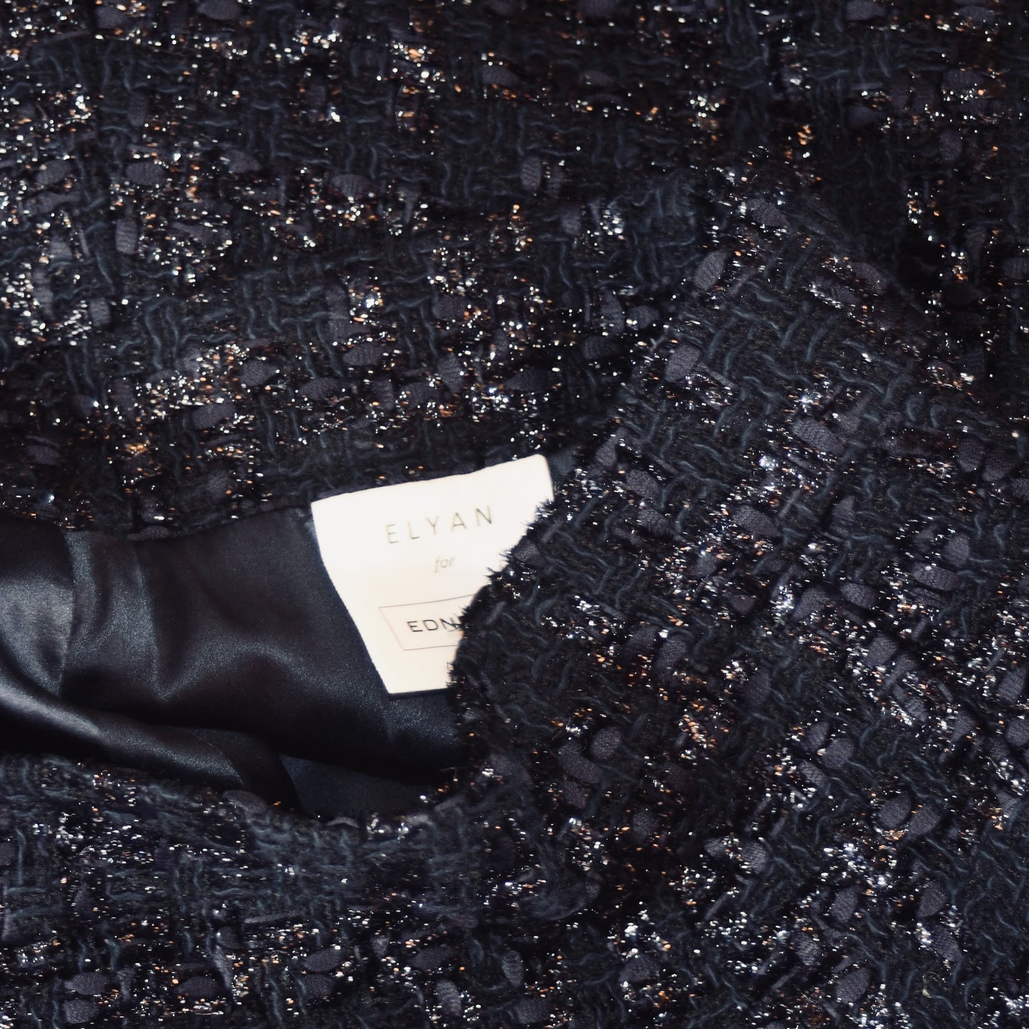 Close-up of the textured, dark, sparkly bouclé fabric from EdNerat’s The Waisted A-Line Skirt in Boucle. The smooth black lining contrasts with the textured outer material. Visible clothing label reads ELYAN for EDNA.