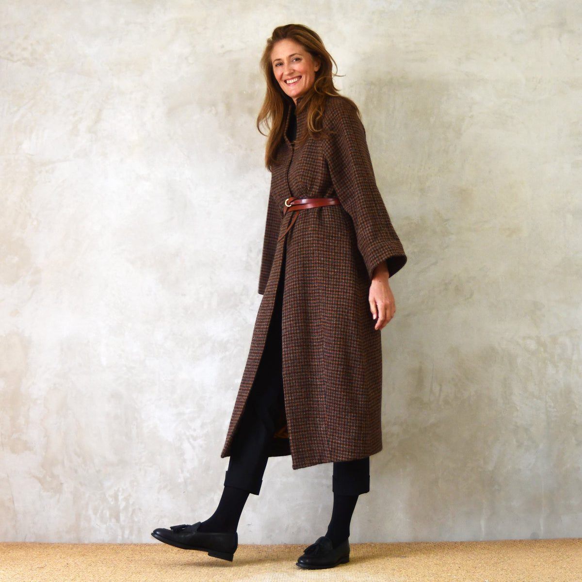 A woman smiles while walking, wearing a long brown checkered coat with a belt, black pants, and black loafers, against a light textured wall background.