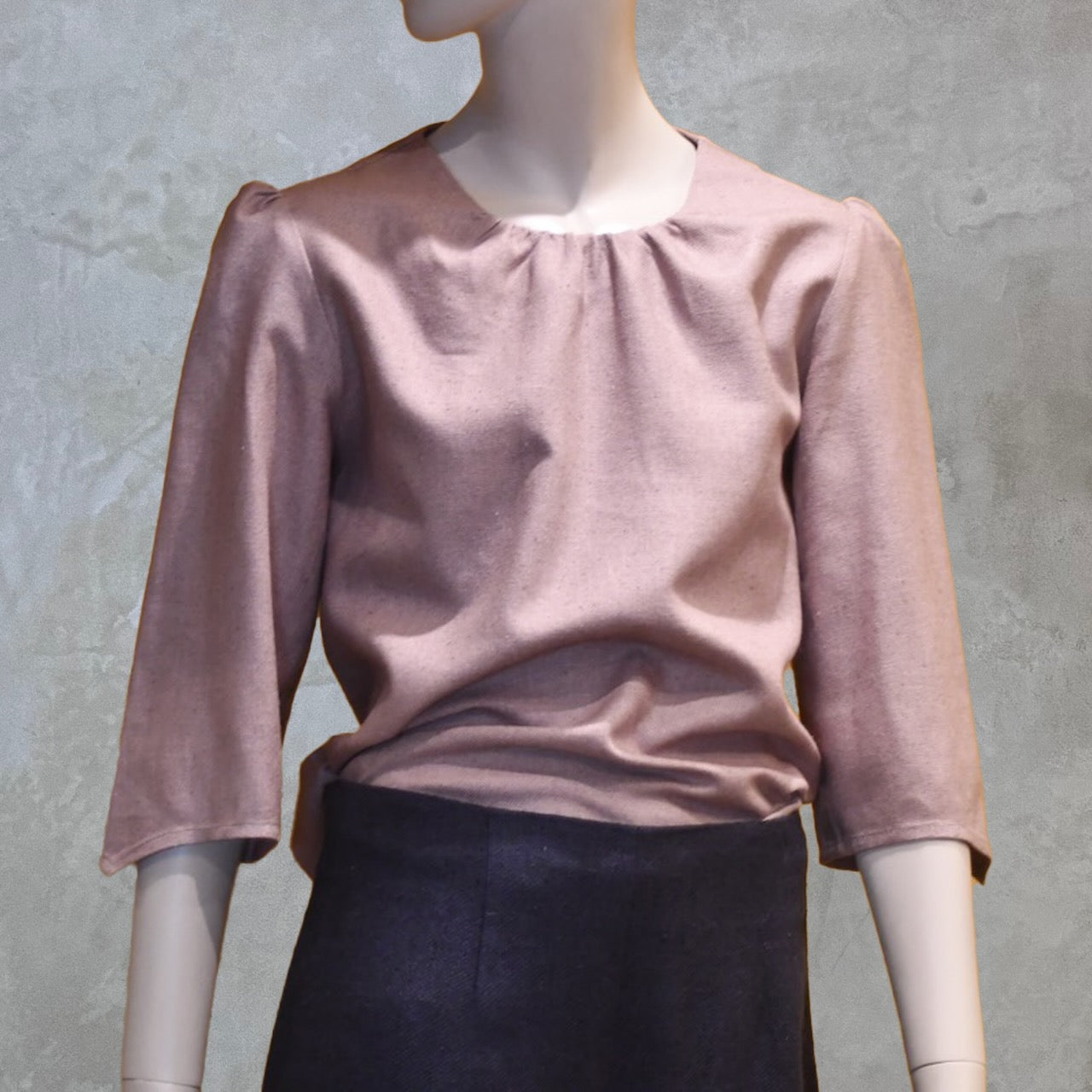 A mannequin dressed in a light pink long-sleeve blouse with a round neckline and dark high-waisted skirt, set against a neutral textured background.