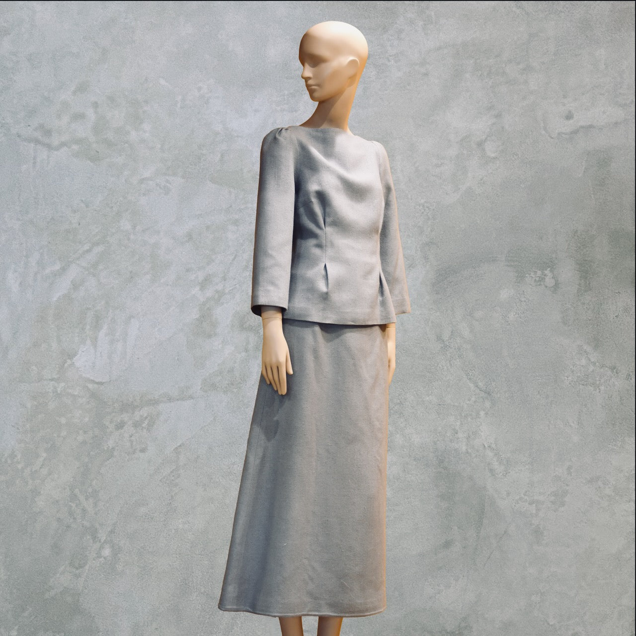 A mannequin dressed in a minimalist, light gray long-sleeve top and matching ankle-length skirt, standing against a textured gray background.