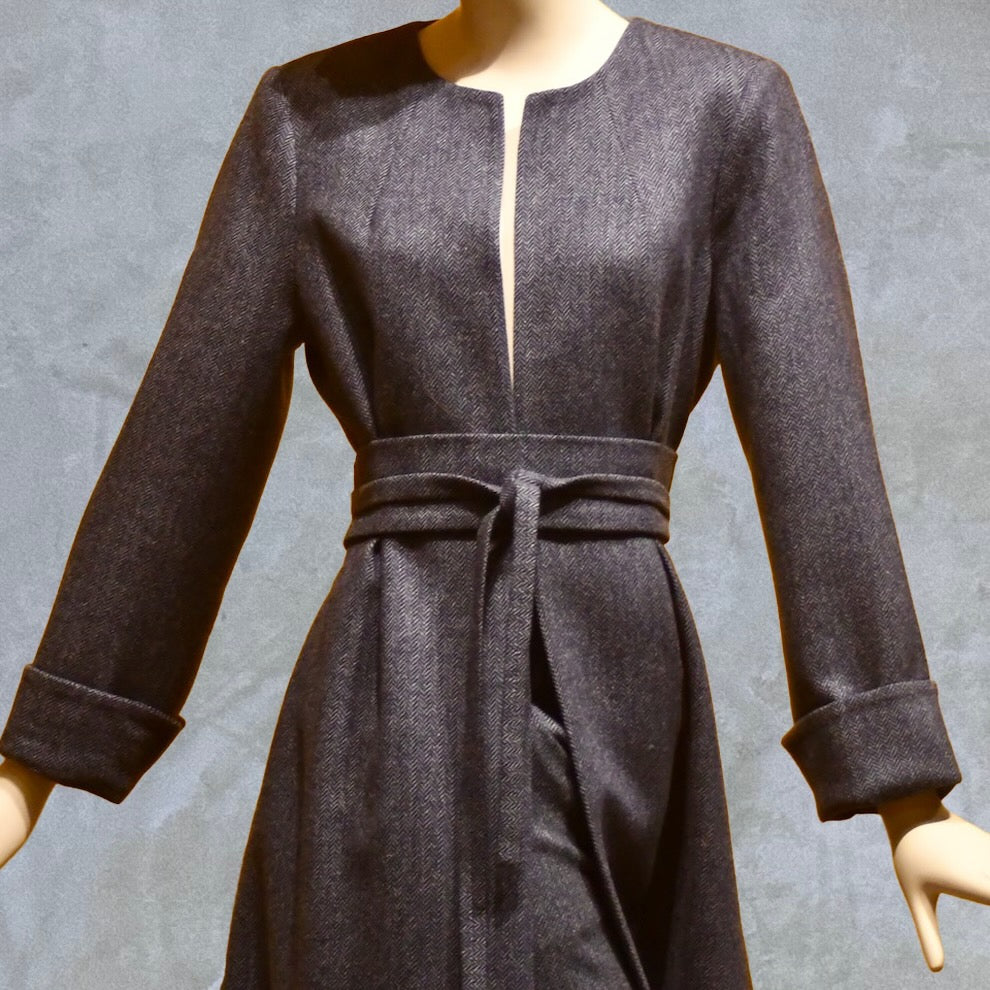 A mannequin wears a long-sleeved, dark gray dress with a front slit, no collar, cuffed sleeves, and a matching waist belt, set against a plain gray background.