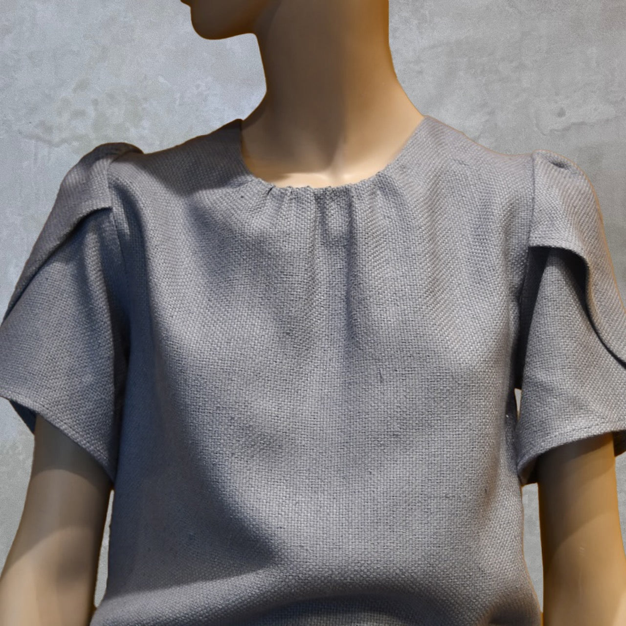 A mannequin wearing a light gray, short-sleeved textured top with gathered neckline and decorative shoulder details, posed against a plain gray background.
