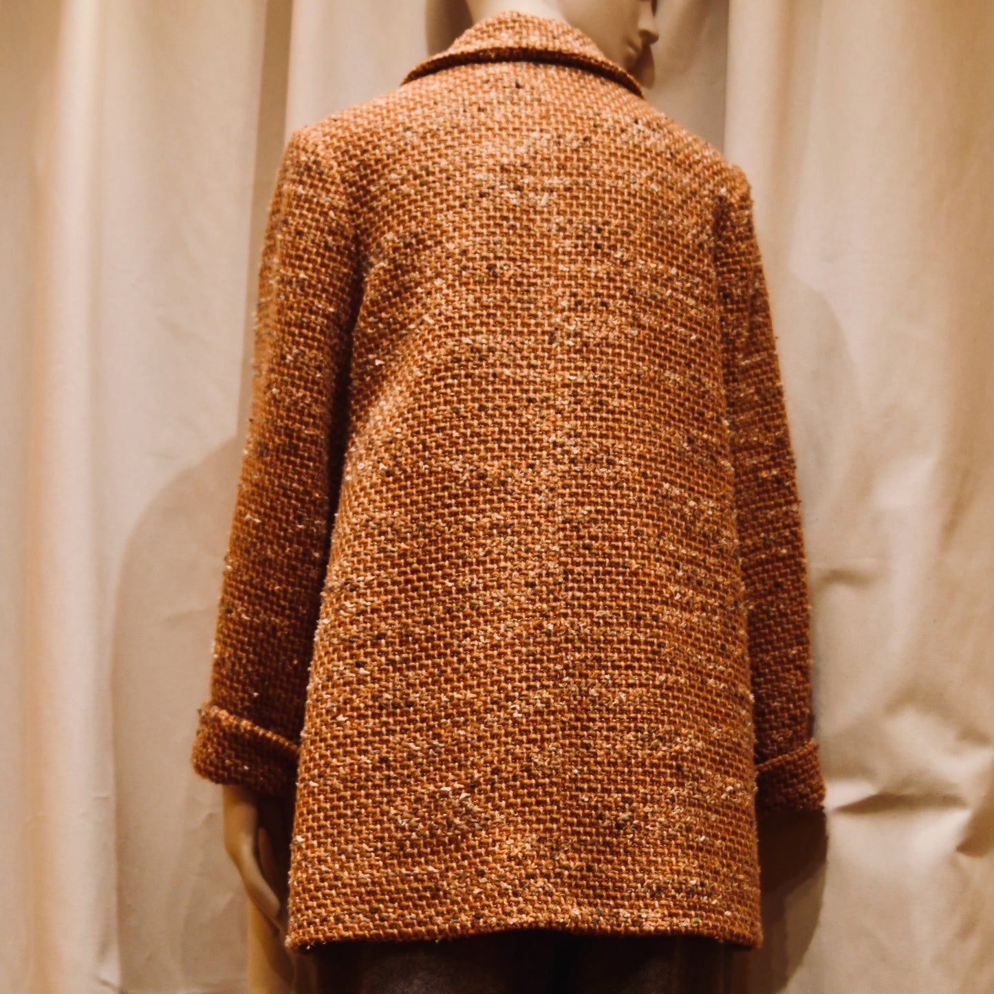 A mannequin stands before beige curtains wearing EdNerats 10 – The Angus Jacket in Burnt Orange Wool Mix Bouclé, shown from the back. The coat features a collar and long sleeves, echoing the signature Angus Jacket style.