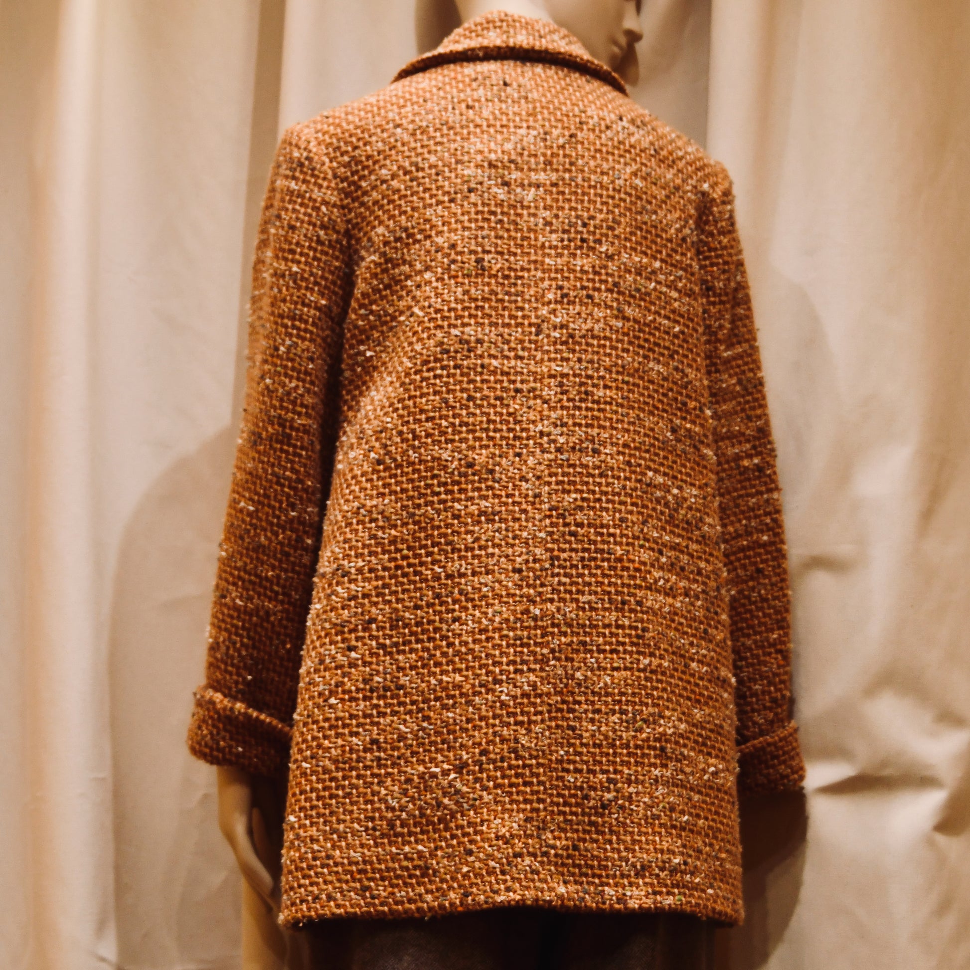 A mannequin stands before beige curtains wearing EdNerats 10 – The Angus Jacket in Burnt Orange Wool Mix Bouclé, shown from the back. The coat features a collar and long sleeves, echoing the signature Angus Jacket style.