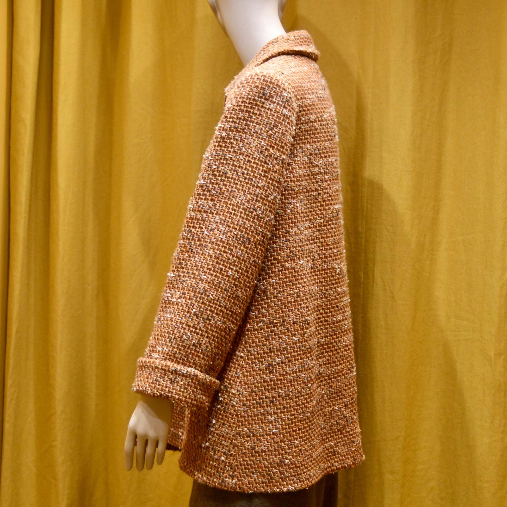A mannequin wears the EdNerat 16 – Angus Jacket in burnt orange wool mix bouclé with rolled-up sleeves, standing against a mustard yellow curtain.