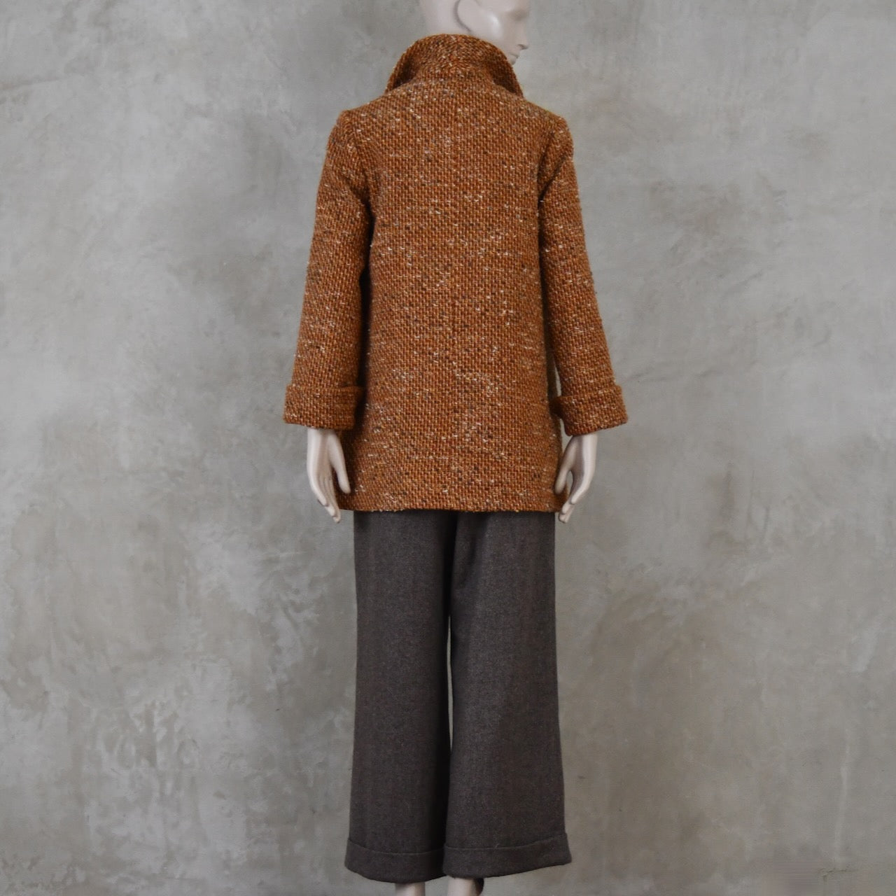 Rear view of Mid length swing jacket in burnt orange Linton Tweeds wool boucle. Handmade in London, this jacket has fold back cuffs, two slanted pockets to the front and a lift up collar. Easy to wear, it is available in sizes 6 to 20 and bespoke sizing. Worn over Fawn Herringbone Merino Flannel Trousers with turn up.