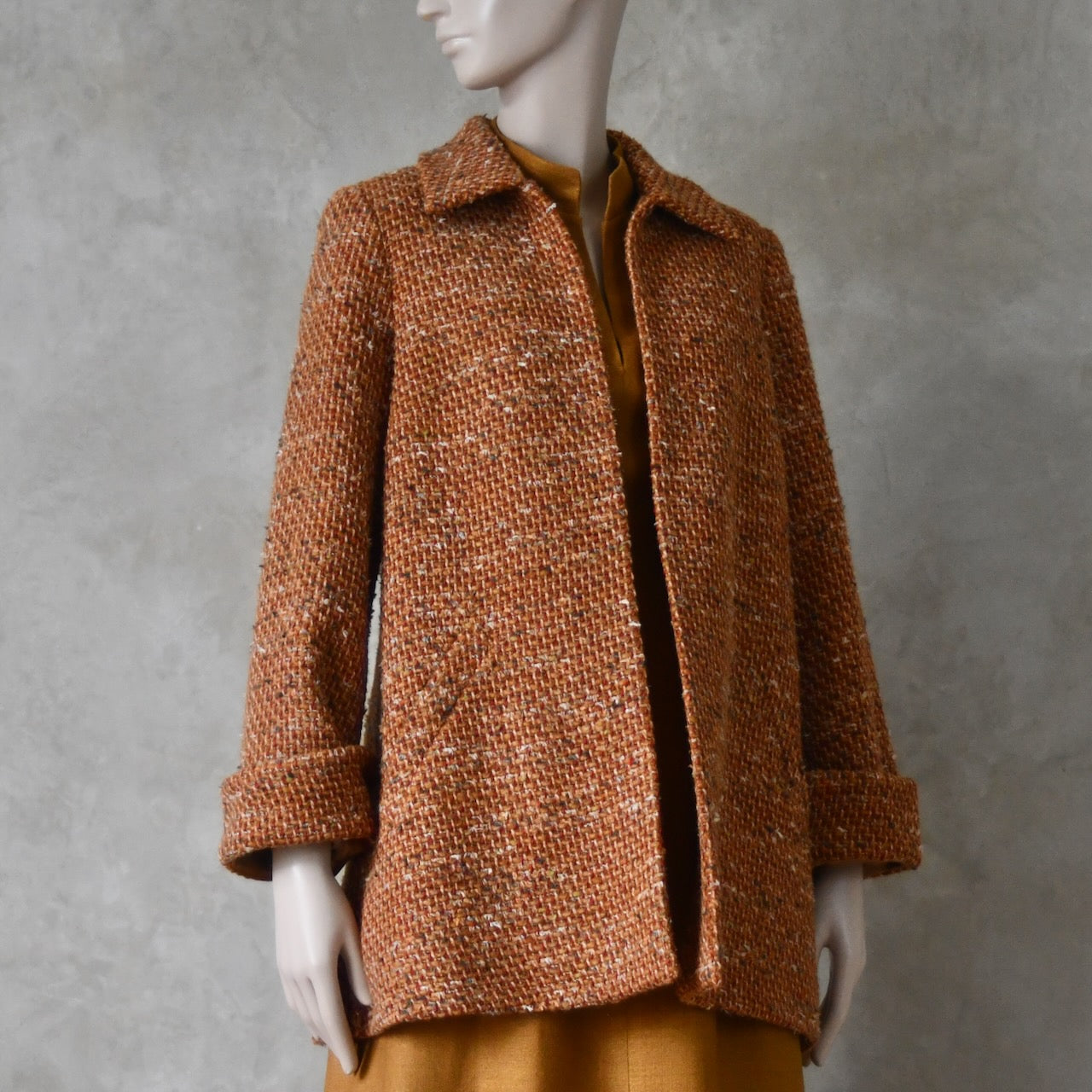 Side open view over tan coloured dress of Mid length swing jacket in burnt orange Linton Tweeds wool boucle. Handmade in London, this jacket has fold back cuffs, two slanted pockets to the front and a lift up collar. Easy to wear, it is available in sizes 6 to 20 and bespoke sizing. 