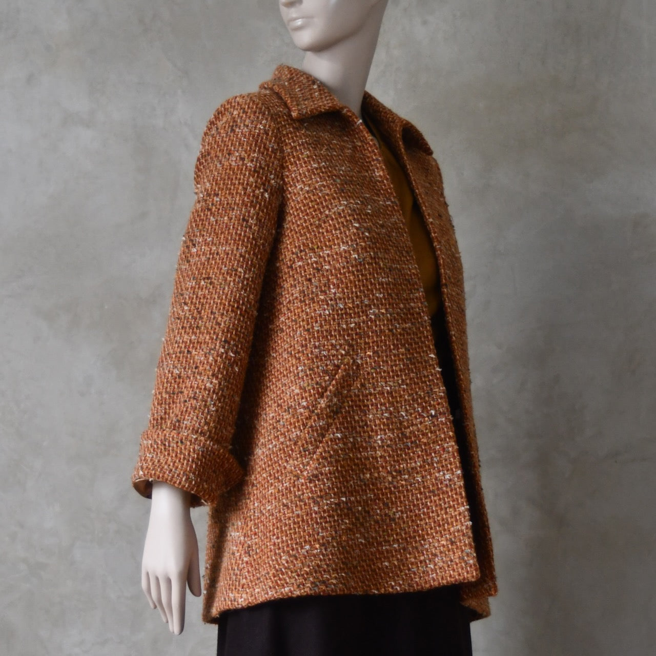 Side open view over brown skirt of Mid length swing jacket in burnt orange Linton Tweeds wool boucle. Handmade in London, this jacket has fold back cuffs, two slanted pockets to the front and a lift up collar. Easy to wear, it is available in sizes 6 to 20 and bespoke sizing. 