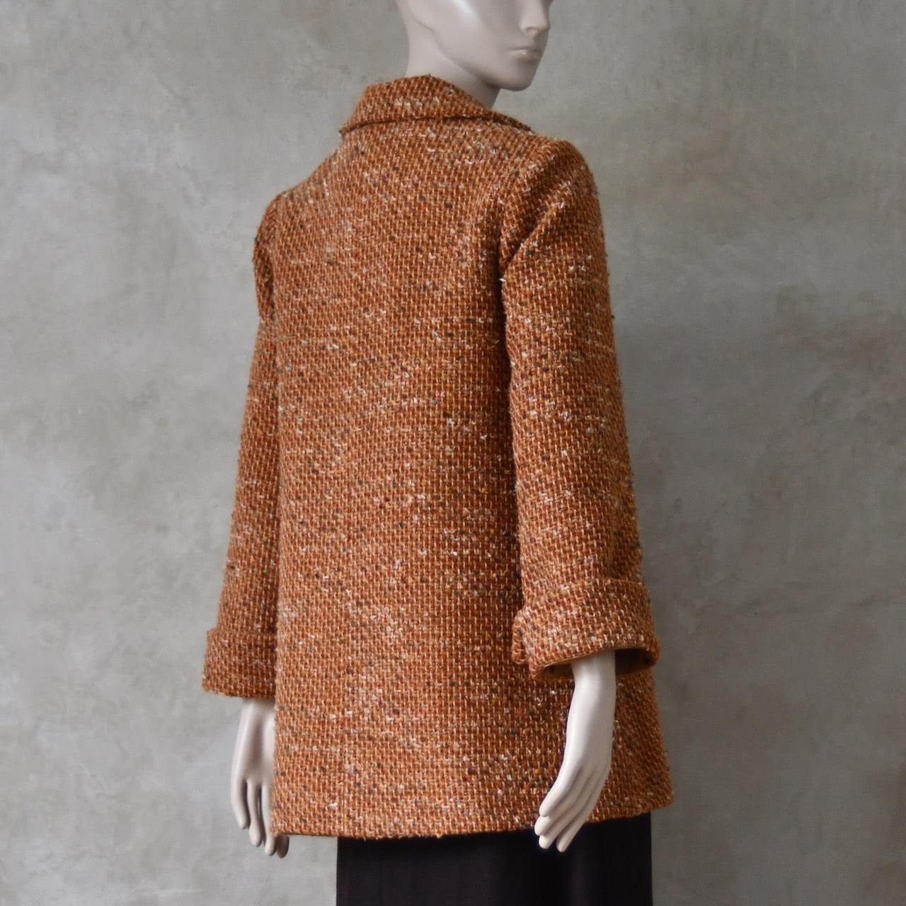 Angled rear view of Mid length swing jacket in burnt orange Linton Tweeds wool boucle. Handmade in London, this jacket has fold back cuffs, two slanted pockets to the front and a lift up collar. Easy to wear, it is available in sizes 6 to 20 and bespoke sizing. 