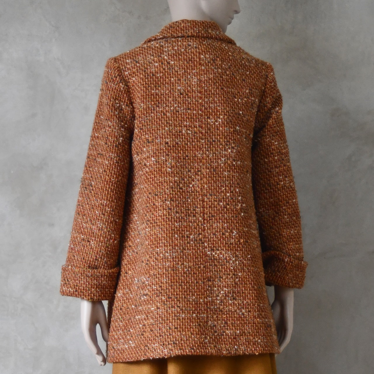Back view of Mid length swing jacket in burnt orange Linton Tweeds wool boucle. Handmade in London, this jacket has fold back cuffs, two slanted pockets to the front and a lift up collar. Easy to wear, it is available in sizes 6 to 20 and bespoke sizing. 