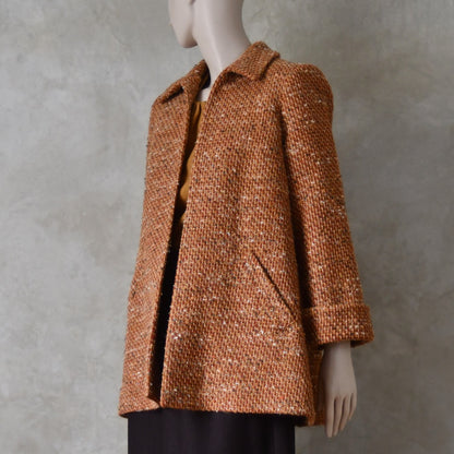 Turning shot of Mid length swing jacket in burnt orange Linton Tweeds wool boucle. Handmade in London, this jacket has fold back cuffs, two slanted pockets to the front and a lift up collar. Easy to wear, it is available in sizes 6 to 20 and bespoke sizing. 