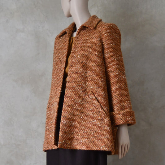 Turning shot of Mid length swing jacket in burnt orange Linton Tweeds wool boucle. Handmade in London, this jacket has fold back cuffs, two slanted pockets to the front and a lift up collar. Easy to wear, it is available in sizes 6 to 20 and bespoke sizing. 