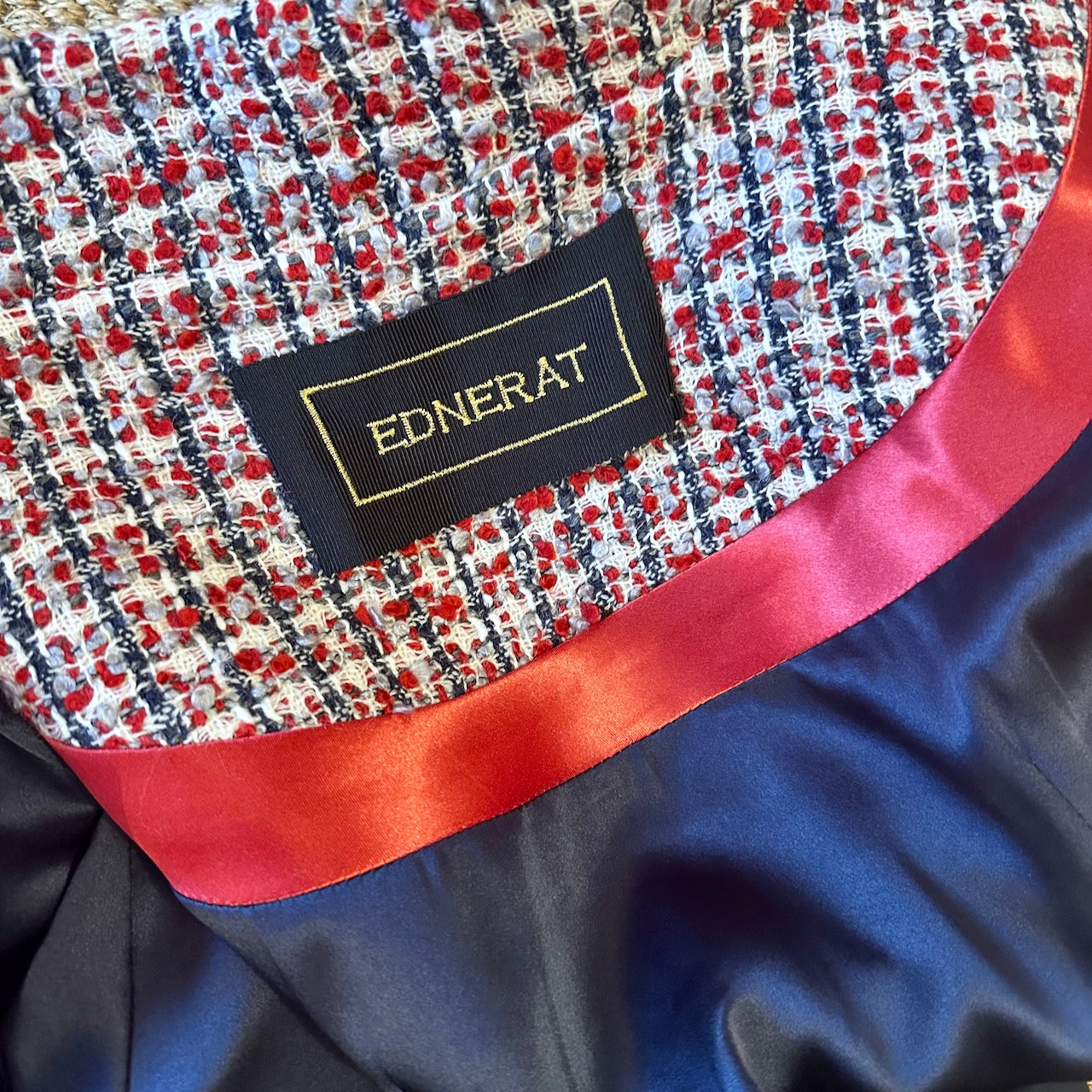 A close-up of an EdNerat label sewn onto red, ecru, and navy wool bouclé fabric with shiny red trim and navy satin—details from The A-Line Coat in Red and Ecru and Navy Wool Boucle from Linton Tweeds.  Handmade in Britain. 