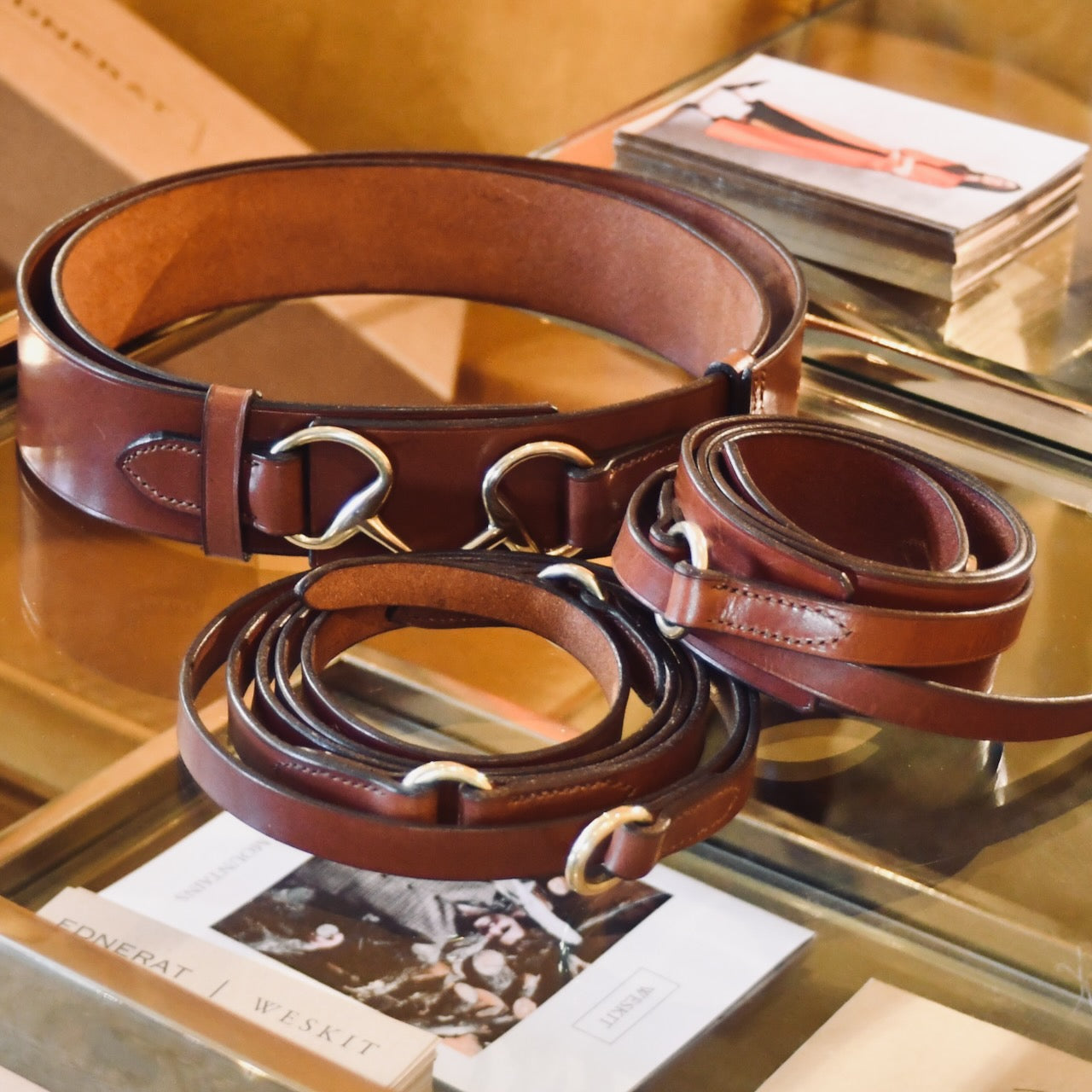 A close-up of several hand-made brown leather belts with silver buckles, neatly coiled and displayed on a glass counter with books and magazines beneath.
