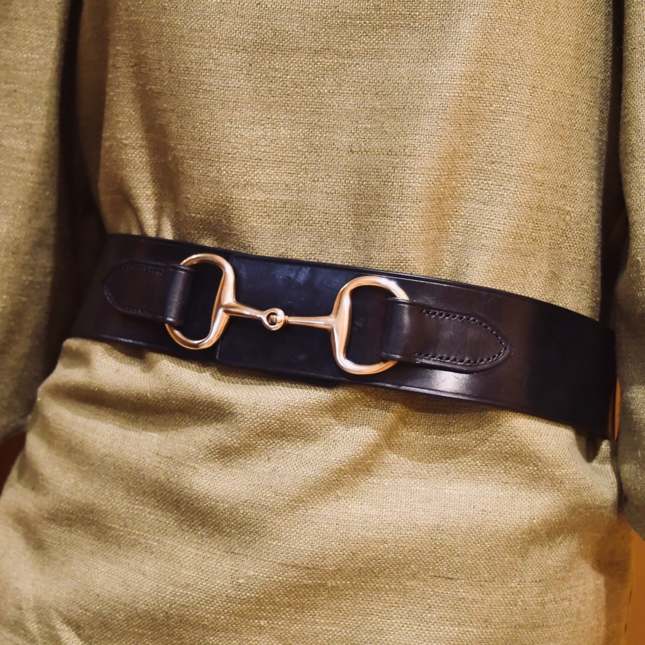 A close-up shows a person wearing the EDNERAT Black Adjustable Bit Belt 80-95 cm, crafted from British saddle leather with solid brass fittings, over a textured olive-green garment.