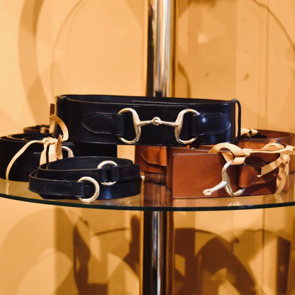 A glass shelf displays the EDNERAT Black Adjustable Bit Belts 80-95 cm, crafted from British saddle leather with solid brass fittings and shiny buckles, arranged neatly, some accented with tan ribbons. Handmade in Britain.
