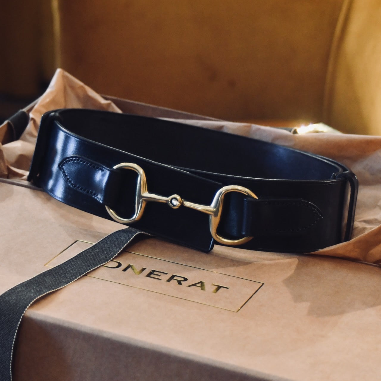 A Black Adjustable Bit Belt 80-95 cm by EDNERAT, made from British saddle leather with a gold horse bit buckle, is displayed on a light brown box with tissue paper. The box bears the EDNERAT logo in gold letters.