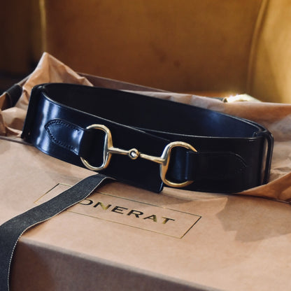 A Black Adjustable Bit Belt 80-95 cm by EDNERAT, made from British saddle leather with a gold horse bit buckle, is displayed on a light brown box with tissue paper. The box bears the EDNERAT logo in gold letters.