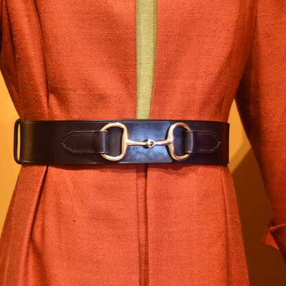 A close-up of a person wearing a rust-colored coat with the EDNERAT Black Adjustable Bit Belt 80-95 cm, crafted from British saddle leather and featuring a silver horse bit buckle at the waist.