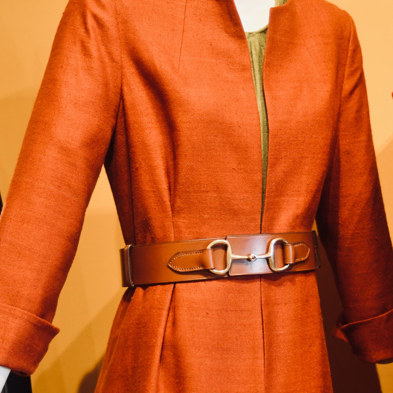 A mannequin wears a rust coloured silk coat with cuffed sleeves, styled with the EDNERAT Hazel Adjustable Bit Belt 95-110 cm, featuring solid brass fittings. Handmade in Britain.