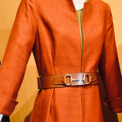 A mannequin wears a rust coloured silk coat with cuffed sleeves, styled with the EDNERAT Hazel Adjustable Bit Belt 95-110 cm, featuring solid brass fittings. Handmade in Britain.