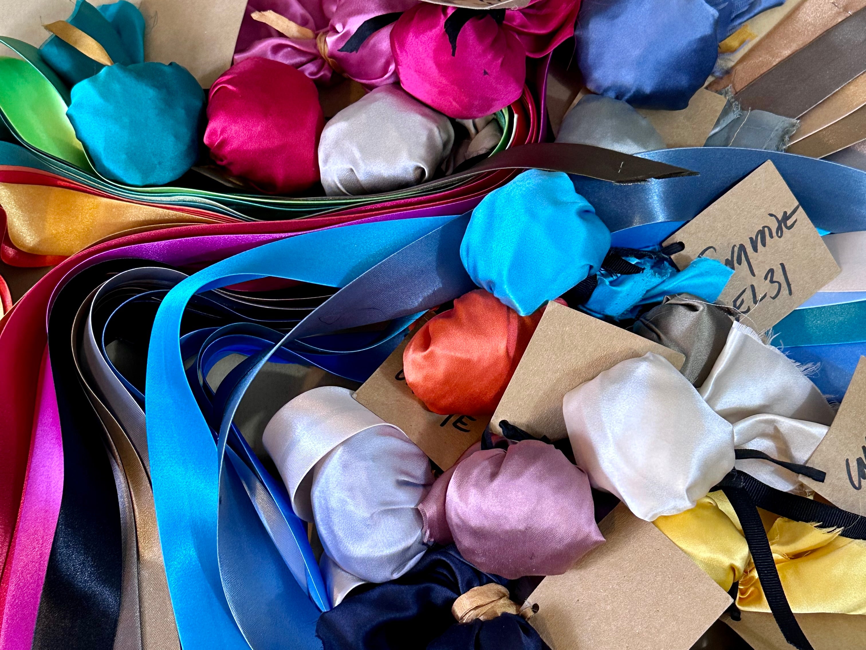 An assortment of colorful, British-made fabric swatches and satin ribbons, with tailored fabric samples tied to brown tags, displayed in a scattered arrangement.