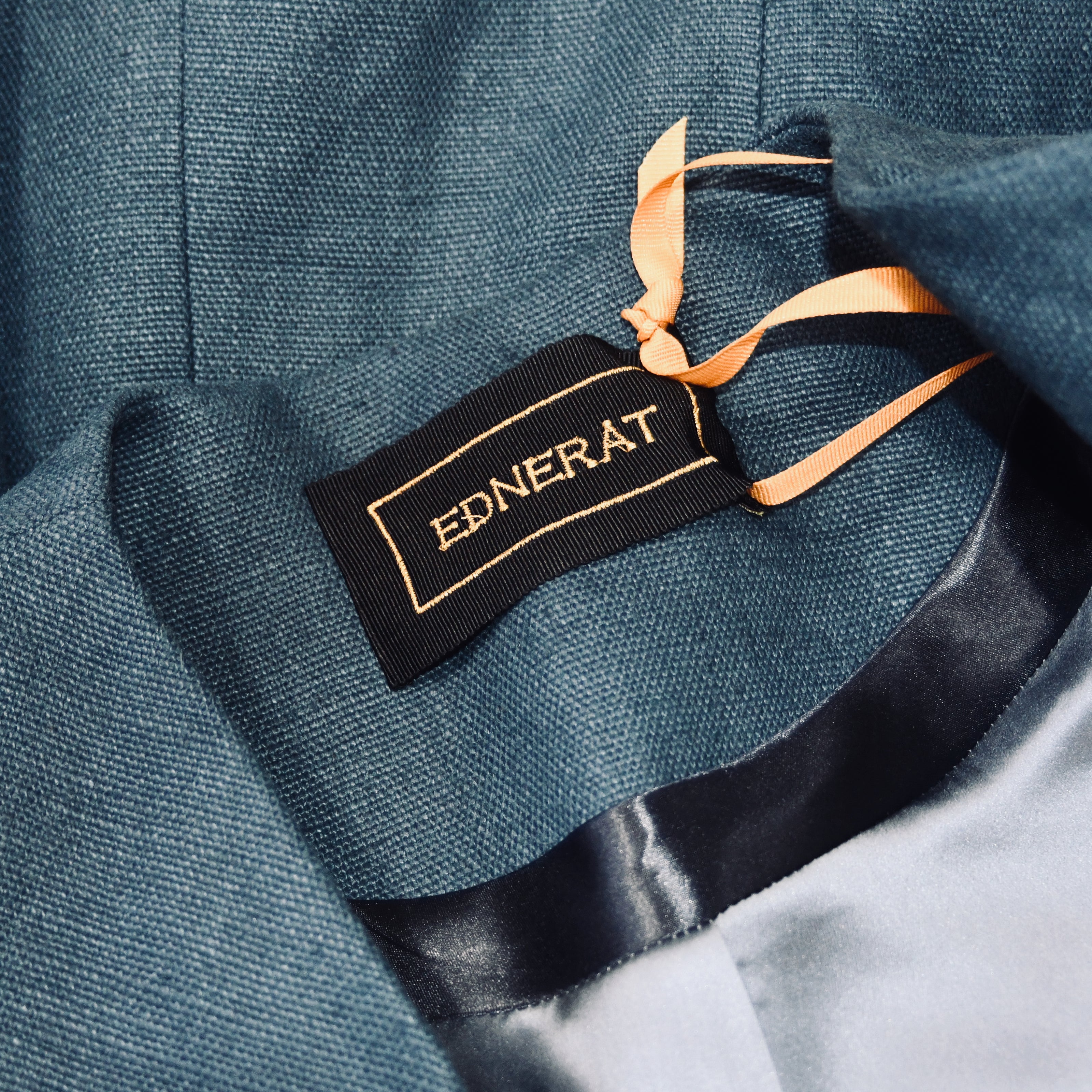 A close-up of a blue blazer’s inner label, reading EDNERAT in gold on black fabric, highlights two attached orange ribbons and a glimpse of the light gray satin lining, showcasing British-made bespoke tailored detail.