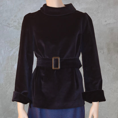 A mannequin models EdNerat’s The Tailored Tunic Top Blouse in Velvet with long sleeves and a wide belt, paired with a blue skirt, against a gray textured background.