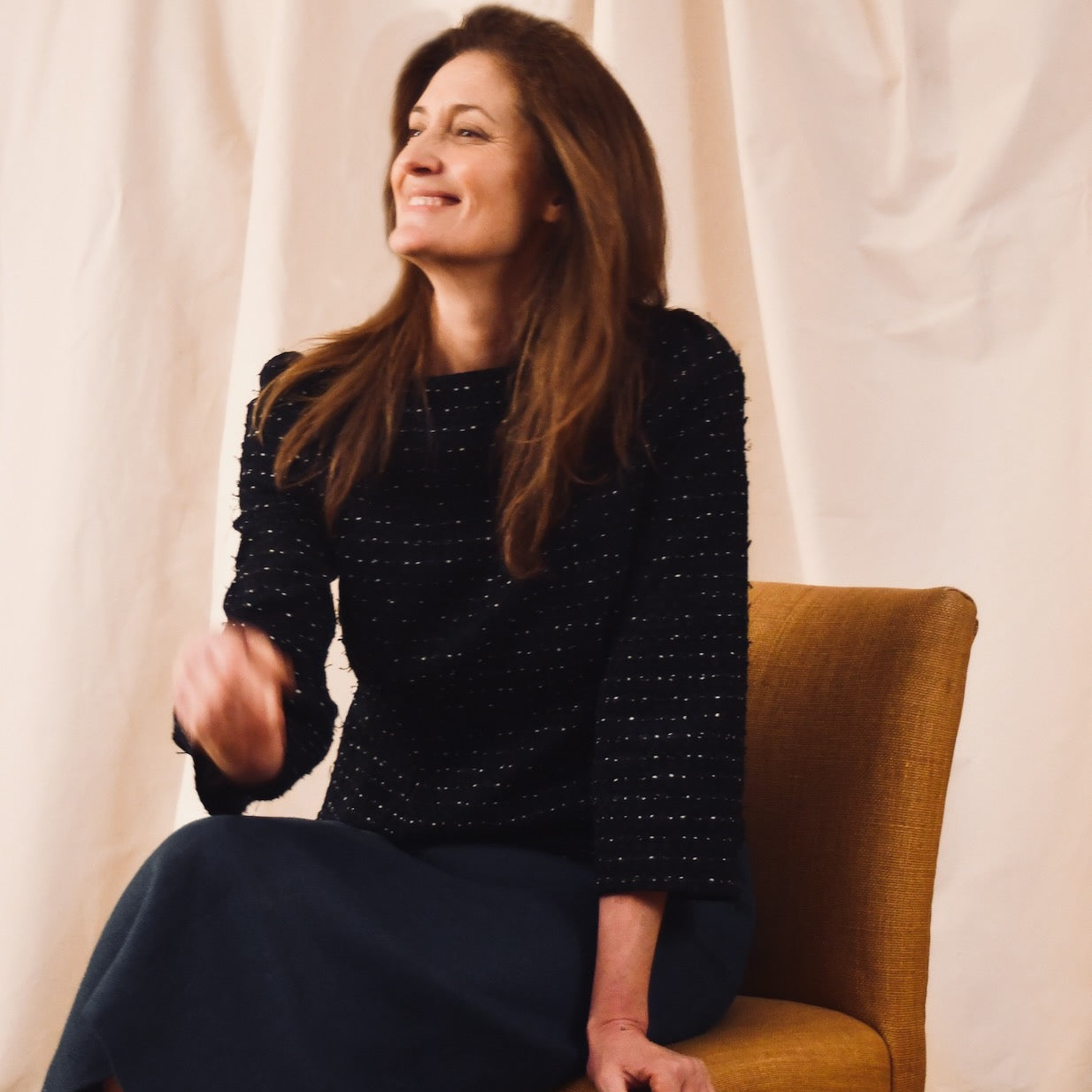 A woman with long brown hair, wearing a bespoke black textured top and dark skirt, sits on a mustard-colored chair, smiling and looking to the side against a neutral fabric backdrop.