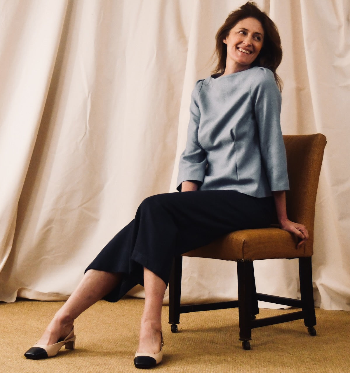 A woman with brown hair sits sideways on a brown chair, smiling and looking to her left. She wears a tailored light blue top, dark pants, and beige shoes with black tips. The background is a cream-colored curtain.