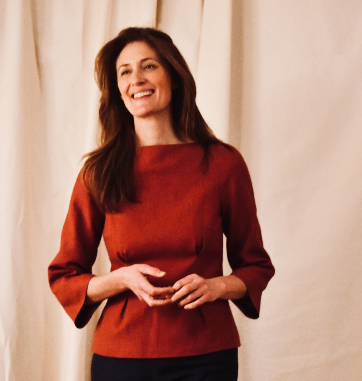 A woman with long brown hair, wearing a tailored rust-colored top with three-quarter sleeves, stands smiling against a beige fabric backdrop. She holds her hands loosely together in front of her.