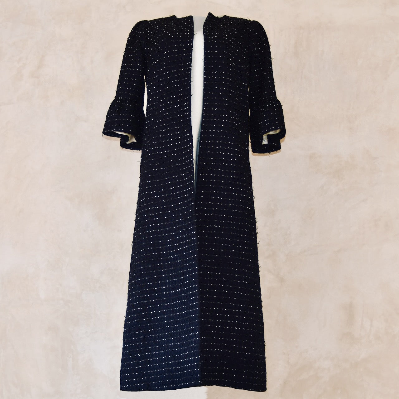 The EdNerat 14 – The Brett Coat in navy, black, and white cotton boucle features a subtle dotted pattern and three-quarter bell sleeves, displayed against a light beige textured background.