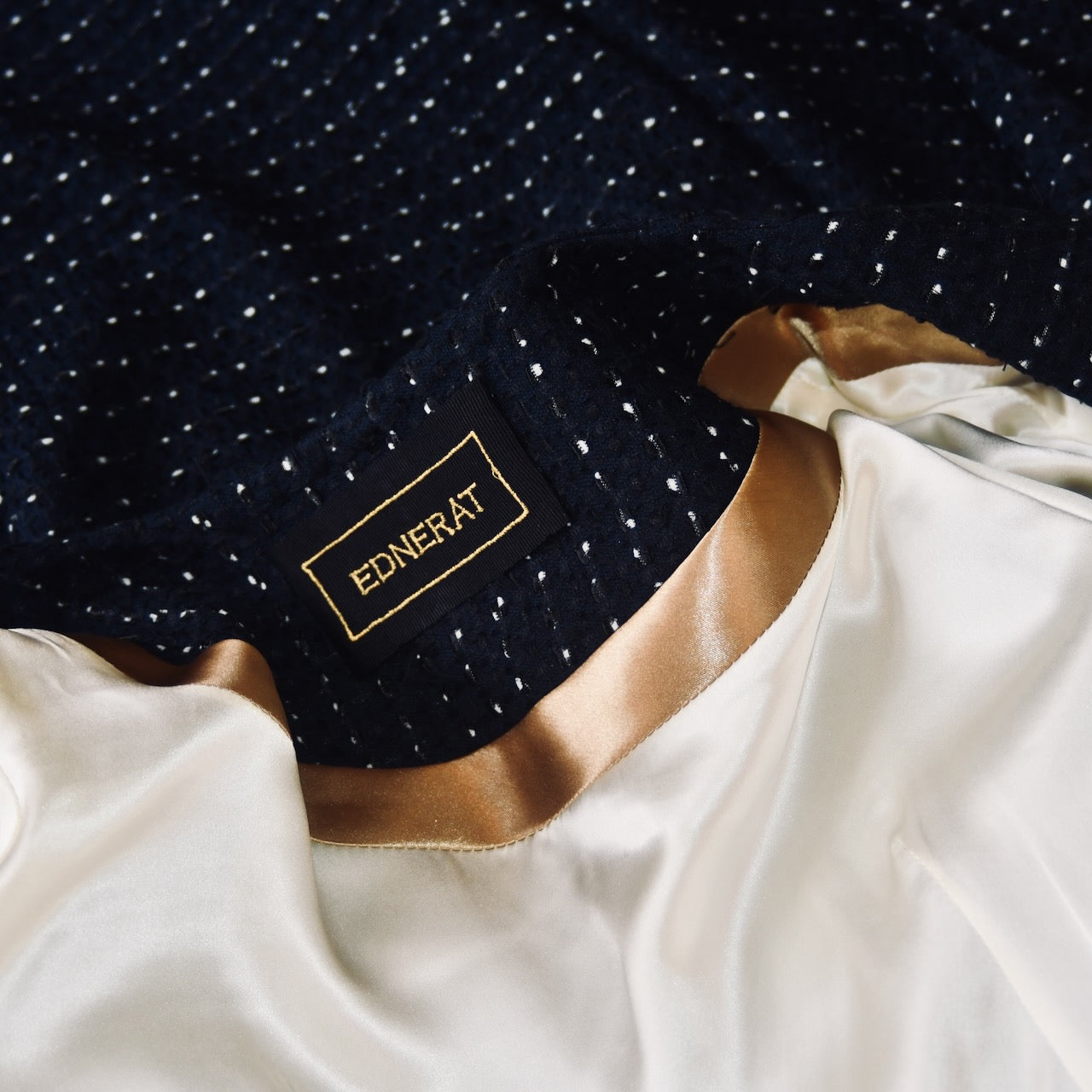 A close-up of an EdNerat label sewn onto navy, black, and white cotton boucle—part of the 14 – The Brett Coat—next to cream and gold satin lining; perfect fabric details for a chic summer look.