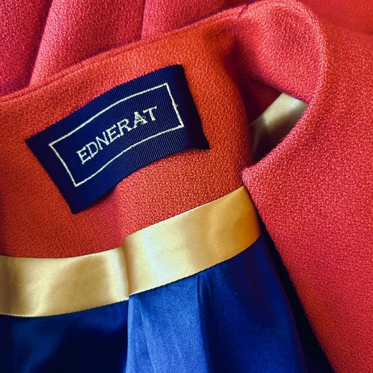 A close-up of The Brett Coat in Burnt Orange Wool Crepe by EdNerat shows the red fabric with gold trim, blue satin lining, and a black silk label with gold embroidery.
