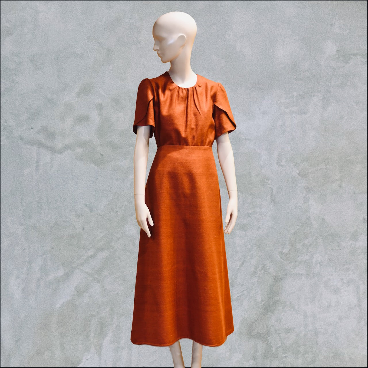A Tulip Sleeve Blouse in Burnt Brick Silk Matka, posed against a textured gray background. Handmade in Britain. 