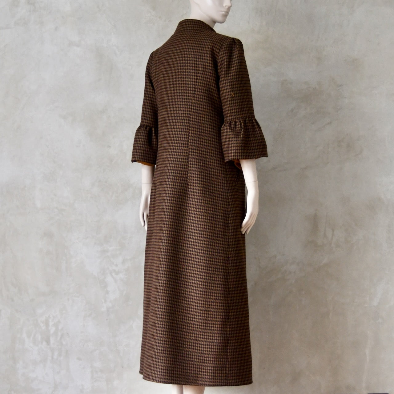 Angled back view of Bespoke Houndstooth patterned Merino Flannel Coat by EdNerat. Handmade in London, this A-Line longer length coat has gathered details at the shoulders, bell sleeves and is lined in pure silk. Woven in Somerset  by Fox Brothers and Handmade in London. 