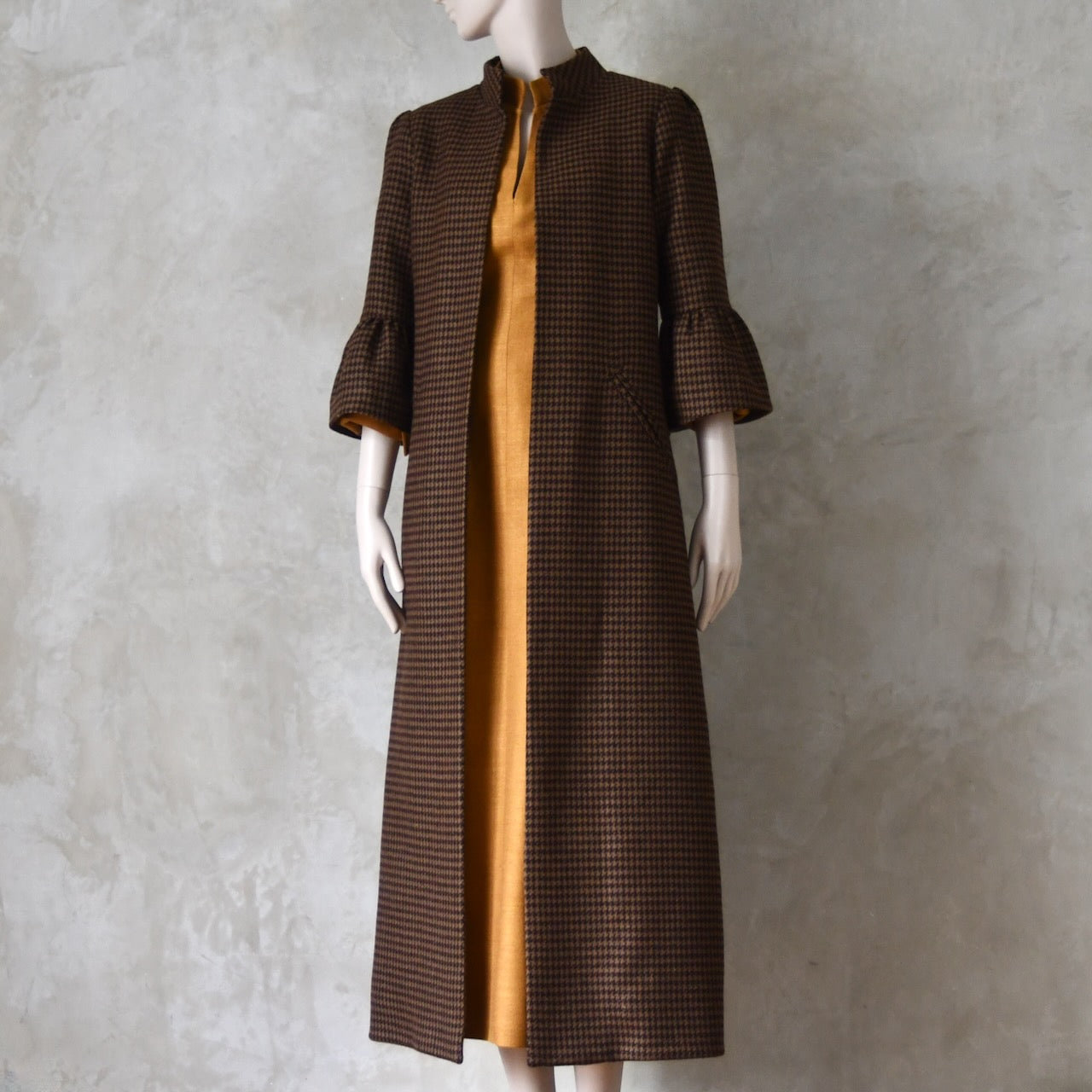 Bespoke Houndstooth patterned Merino Flannel Coat by EdNerat. Handmade in London, this A-Line longer length coat has gathered details at the shoulders, bell sleeves and is lined in pure silk. Woven in Somerset  by Fox Brothers and Handmade in London. Shown  at an angle over Silk Waisted Dress.