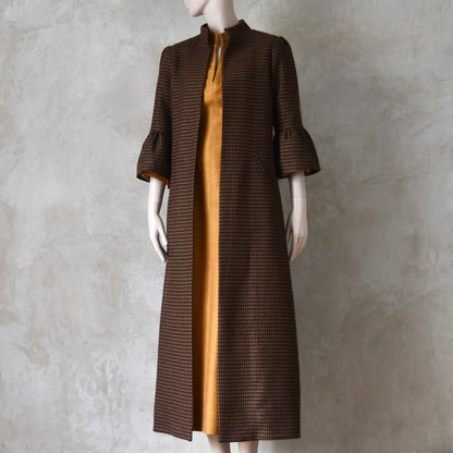 Bespoke Houndstooth patterned Merino Flannel Coat by EdNerat. Handmade in London, this A-Line longer length coat has gathered details at the shoulders, bell sleeves and is lined in pure silk. Woven in Somerset  by Fox Brothers and Handmade in London. Shown  at an angle over Silk Waisted Dress.