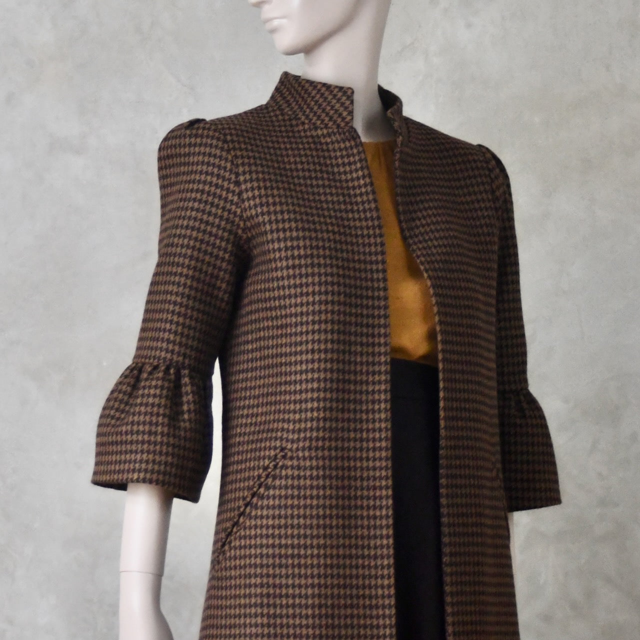 Bespoke Houndstooth patterned Merino Flannel Coat by EdNerat. Handmade in London, this A-Line longer length coat has gathered details at the shoulders, bell sleeves and is lined in pure silk. Woven in Somerset  by Fox Brothers and Handmade in London. Shown over Tan Top and Brown A Line Skirt.