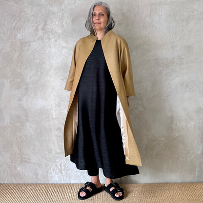 A woman with long gray hair stands indoors against a textured light gray wall, wearing a black dress, black sandals, and the Silk Matka Cocoon Coat by EdNerat. She gazes slightly to the side with a neutral expression.
