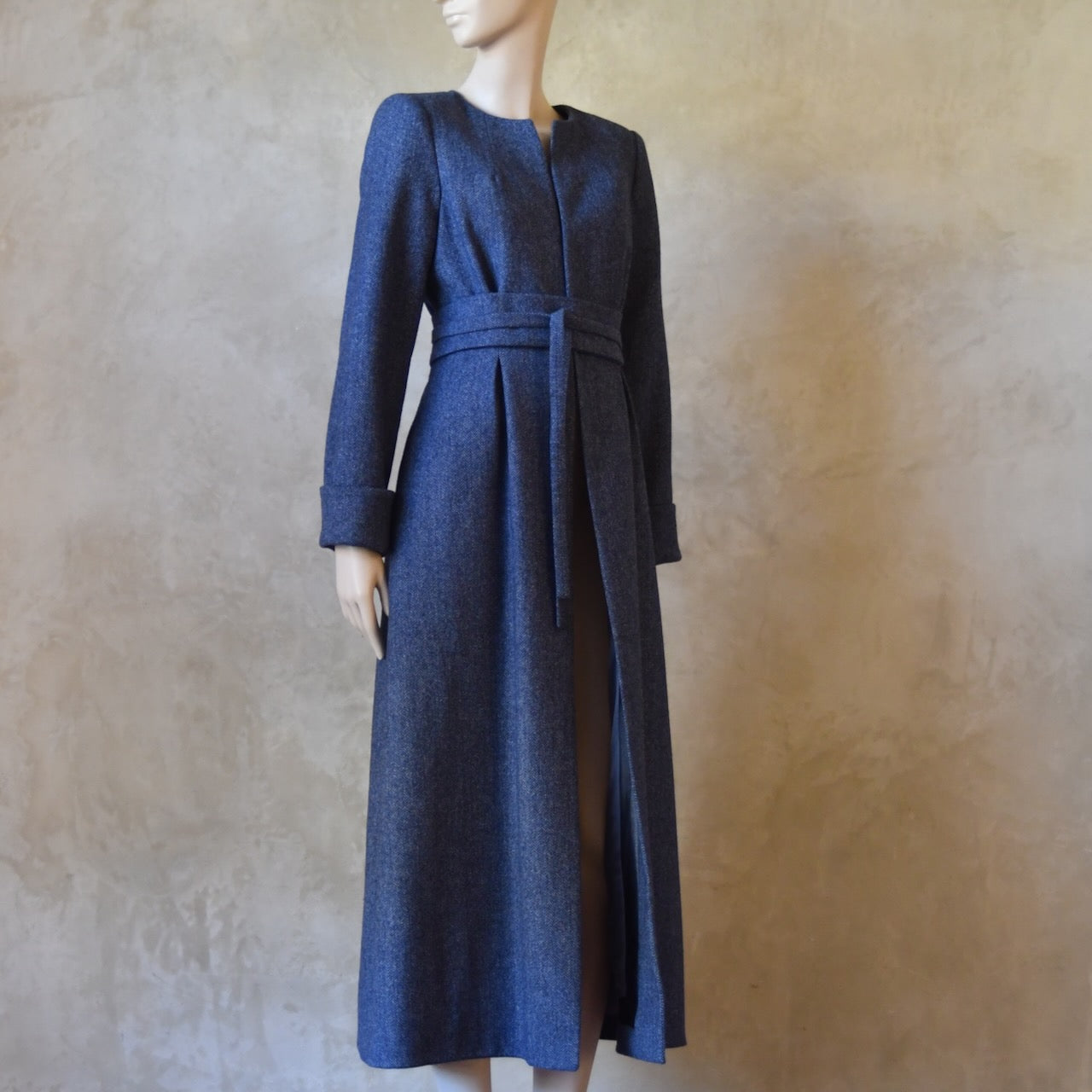 Side Angled view of Bespoke Belted Blue Tweed Coat, Handmade in London. Denim Blue herringbone tweed dress coat, ideal for winter weddings, race fixtures and other formalwear occasions. Fully lined in pure silk, with optional Nehru collar, fold back cuffs and self-fabric wrap around belt.
Longer or calf length coat.