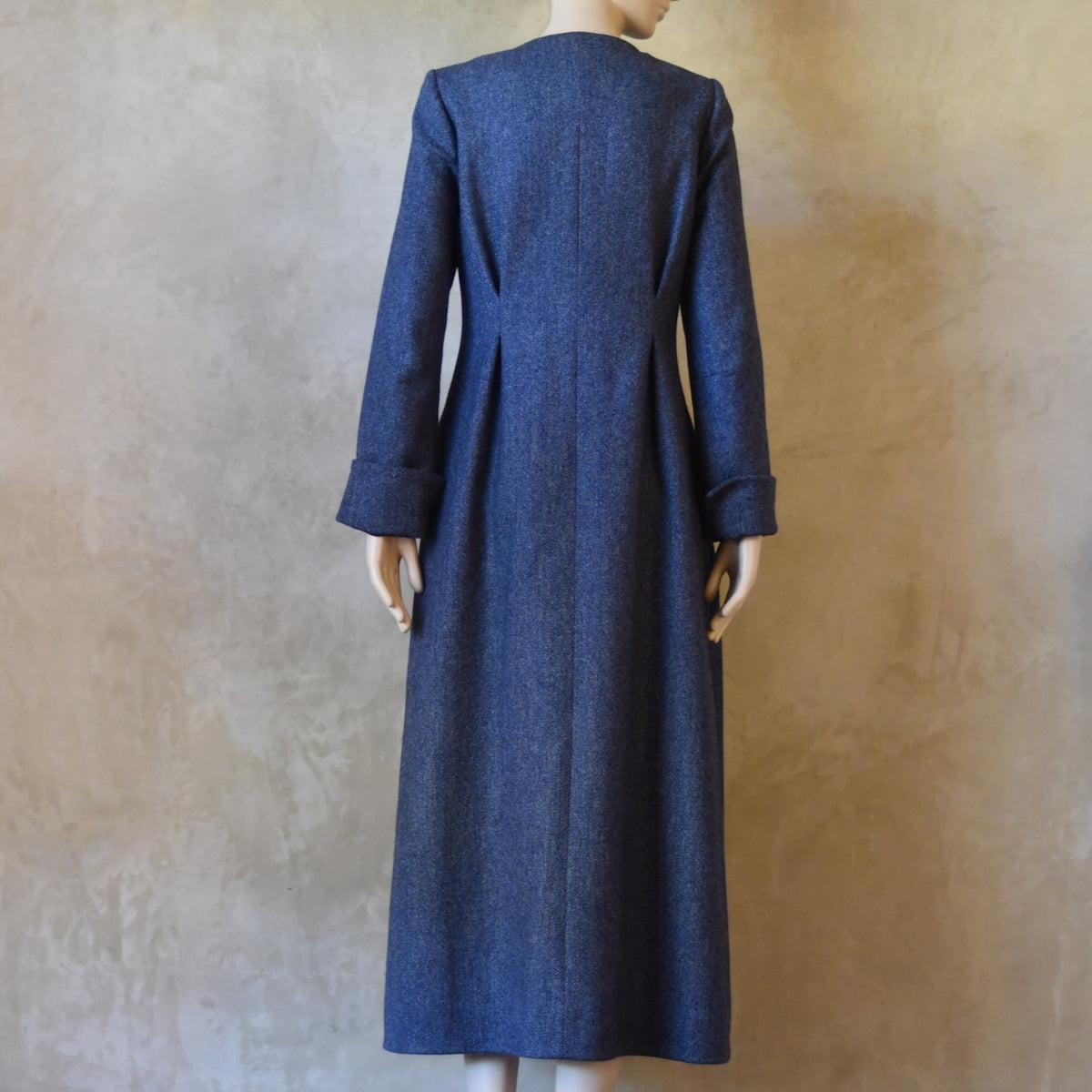 Rear view of Bespoke Hourglass Tweed Coat, Handmade in London. Denim Blue herringbone tweed dress coat, ideal for winter weddings, race fixtures and other formalwear occasions. Fully lined in pure silk, with optional Nehru collar, fold back cuffs and self-fabric wrap around belt.
Longer or calf length coat.