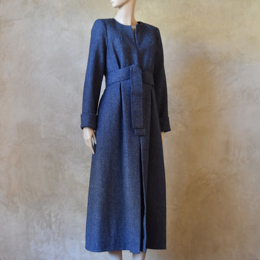 Side Angled view of Bespoke Hourglass Tweed Coat, Handmade in London. Denim Blue herringbone tweed dress coat, ideal for winter weddings, race fixtures and other formalwear occasions. Fully lined in pure silk, with optional Nehru collar, fold back cuffs and self-fabric wrap around belt.
Longer or calf length coat.