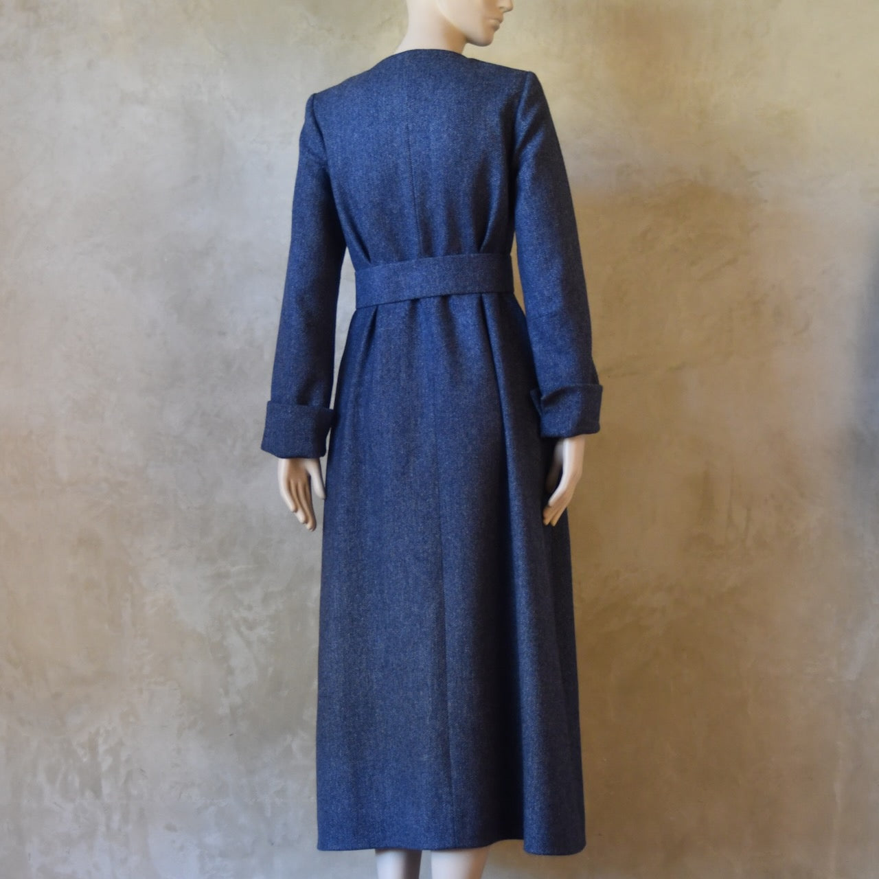 Rear belted view of Bespoke Hourglass Tweed Coat, Handmade in London. Denim Blue herringbone tweed dress coat, ideal for winter weddings, race fixtures and other formalwear occasions. Fully lined in pure silk, with optional Nehru collar, fold back cuffs and self-fabric wrap around belt.
Longer or calf length coat.