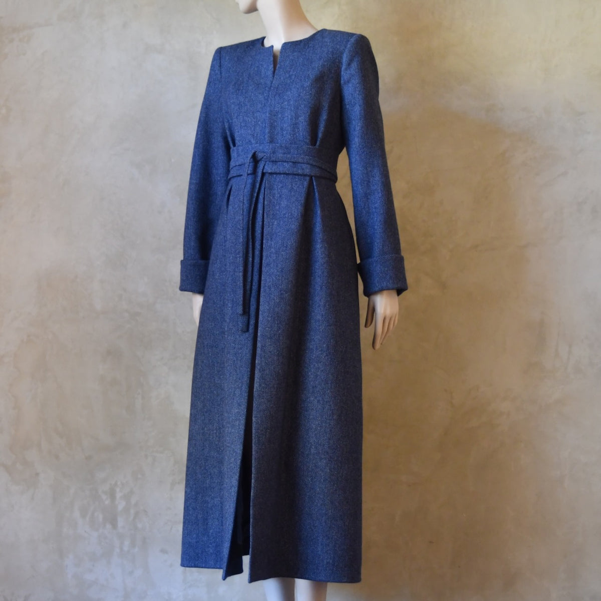 Angled front belted view of Bespoke Hourglass Tweed Coat, Handmade in London. Denim Blue herringbone tweed dress coat, ideal for winter weddings, race fixtures and other formalwear occasions. Fully lined in pure silk, with optional Nehru collar, fold back cuffs and self-fabric wrap around belt.
Longer or calf length coat.